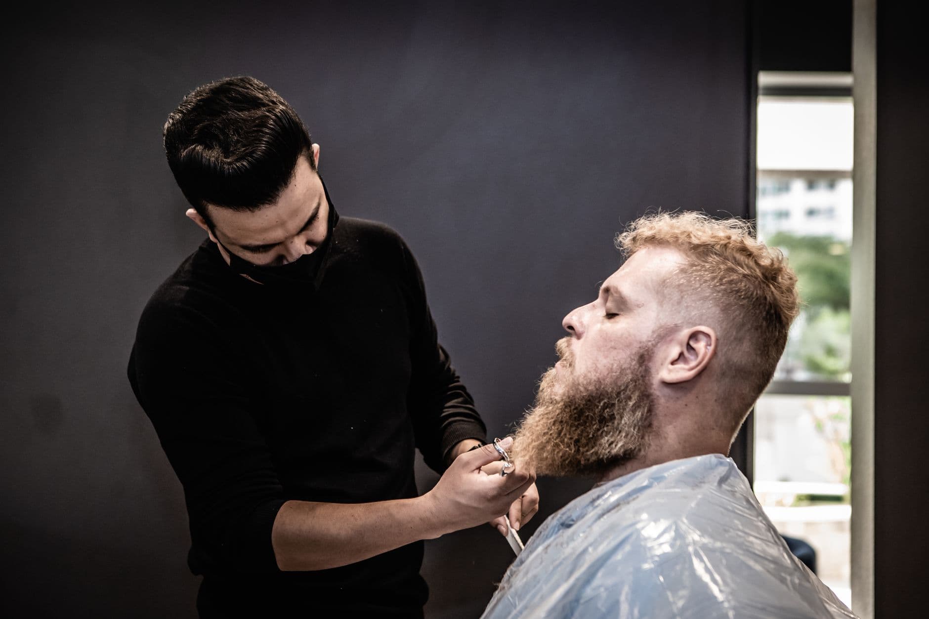 Barbeiro apara a barba de um cliente sentado na cadeira da barbearia.